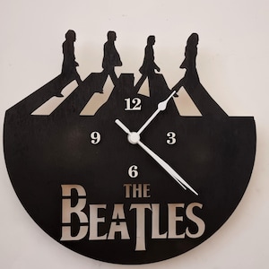 Puede incluir: Reloj de pared de madera negra con la icónica escena del cruce de Abbey Road con cuatro figuras. La esfera del reloj tiene números y manecillas blancas, y las palabras "THE BEATLES" se muestran en la parte inferior.