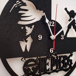 May include: A black and white circular wall clock with a cutout of a character from the anime One Piece. The clock face has white numbers and hands. The character is wearing a straw hat and has a serious expression. The clock is surrounded by a black frame and the words 'One Piece' are written in white letters below the character.