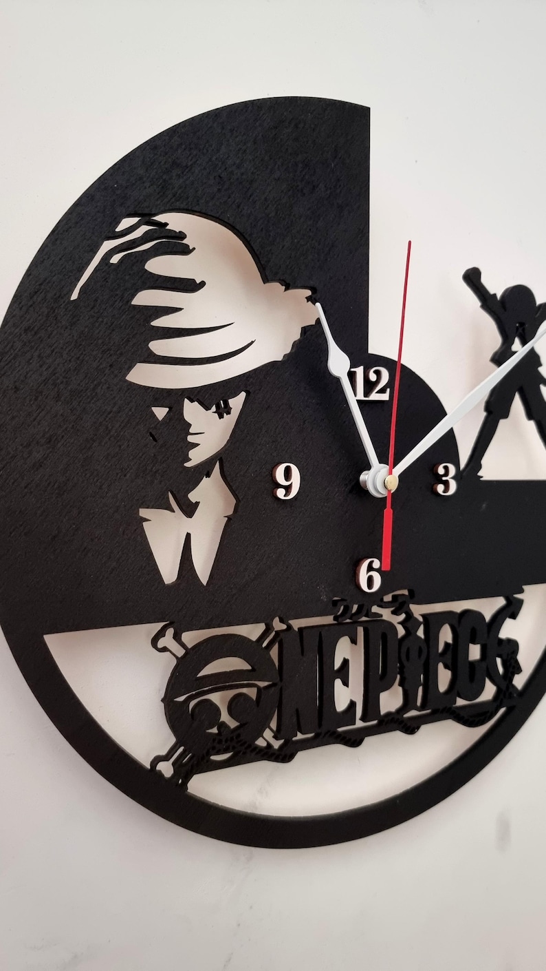 May include: A black and white circular wall clock with a cutout of a character from the anime One Piece. The clock face has white numbers and hands. The character is wearing a straw hat and has a serious expression. The clock is surrounded by a black frame and the words 'One Piece' are written in white letters below the character.