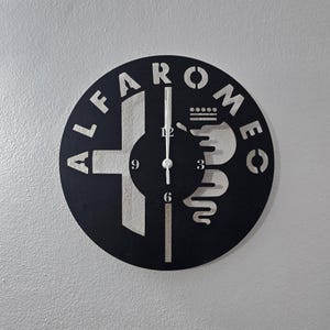Może przedstawiać: Czarny okrągły zegar ścienny z logo Alfa Romeo. Zegar ma białe wskazówki i cyfry, z nazwą marki na obrzeżu. Zegar jest zamontowany na jasnoszarej ścianie.