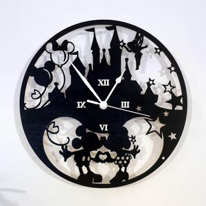 Può includere: Orologio da parete circolare nero con un design a silhouette di un castello, Topolino e Minnie, stelle e una fata. Le lancette dell'orologio sono bianche. Il quadrante ha numeri romani. È un oggetto decorativo.