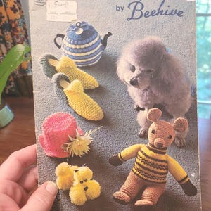 May include: A vintage book cover titled "Bazaar Novelties and Gifts by Beehive" featuring various knitted items. The cover displays a blue and white teapot, slippers, a pink hat, a poodle, a bear, and a bee-themed figure.