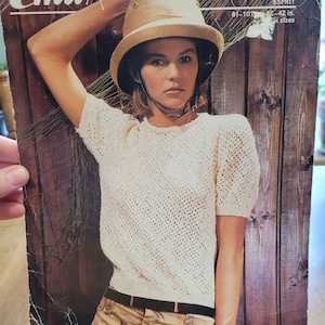 Könnte beinhalten: Ein weißer Häkelpullover mit kurzen Ärmeln und einem braunen Gürtel. Das Model trägt einen braunen Safarihut und eine Khakihose mit Tarnmuster. Das Bild enthält den Text "Emu" und "3755 Esprit 32-42 in. 6 Größen".