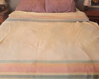 Puede incluir: Una cama blanca con una manta a rayas de color azul claro, rosa y crema. La cama tiene una cabecera de madera con un tapiz de macramé detrás.