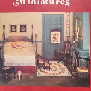 Op de afbeelding: Een rode boekomslag met de titel "Charted Designs For Miniatures" in witte letters. De afbeelding toont een miniatuur slaapkamer met een bed, stoelen en een tapijt. Het boek is onderaan gemarkeerd met "Book 2".