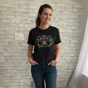 May include: A black t-shirt featuring a colorful floral design with red, blue, and green elements. The shirt is paired with blue jeans and white sneakers. The person is standing in front of a brick wall.