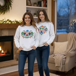 May include: Two people wearing white sweatshirts with colorful floral and rooster designs, paired with blue jeans and white boots. The setting is a cozy room with a fireplace and a neutral-toned rug.