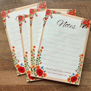 May include: A stack of floral-themed notepads. Each notepad features a cream-colored background with a decorative border of red poppies, orange, and blue flowers. The word "Notes" is written in elegant script, and the bottom of the notepad reads "Small steps today creating big results tomorrow."