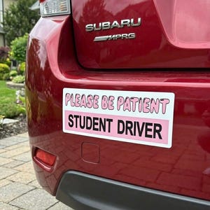 May include: A red Subaru car with a white and pink "PLEASE BE PATIENT STUDENT DRIVER" bumper sticker. The car's taillight and bumper are visible.