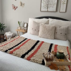 May include: A chunky knit throw blanket with stripes of beige, black, and coral. The blanket is draped over a bed with white linens and several decorative pillows. A wooden tray with a small pumpkin, fall leaves, and a ceramic jar is on the bed in front of the blanket.