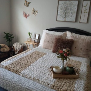 May include: A white and beige chunky knit throw blanket on a bed with white and beige pillows. There are two framed pictures on the wall behind the bed, one with a tree and the other with a plant.