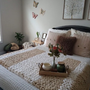 May include: A white and beige knitted throw blanket on a bed with a floral arrangement in a vase and candles on a wooden tray. The bed has a white and beige patterned headboard and a white and beige patterned bedspread. There are three butterfly wall decorations on the wall behind the bed.