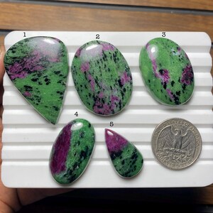 May include: Five polished ruby in zoisite gemstones in various shapes, including oval and teardrop. The stones display a mix of green and pink hues, with a quarter dollar coin for size comparison. The gemstones are set on a white surface.