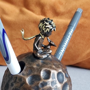 Op de afbeelding: Een bronzen pennenhouder in de vorm van een planeet, met een kleine sculptuur van een zittende figuur. Twee pennen zijn in de houder geplaatst. De achtergrond omvat een oranje kussen en een grijs oppervlak.