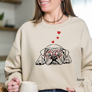 May include: A beige sweatshirt with a black and white illustration of a bulldog wearing red heart-shaped glasses. The bulldog is lying down and there are red hearts floating above it. The text "Sand" is printed on the bottom right corner of the sweatshirt.