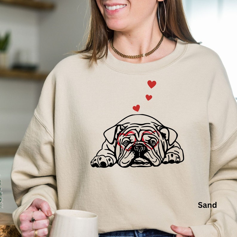 May include: A beige sweatshirt with a black and white illustration of a bulldog wearing red heart-shaped glasses. The bulldog is lying down and there are red hearts floating above it. The text "Sand" is printed on the bottom right corner of the sweatshirt.