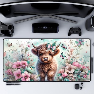 May include: A rectangular mouse pad featuring a watercolor illustration of a Highland cow with a pink and blue bow, surrounded by pink flowers and butterflies. The pad has a black border and is on a white surface.