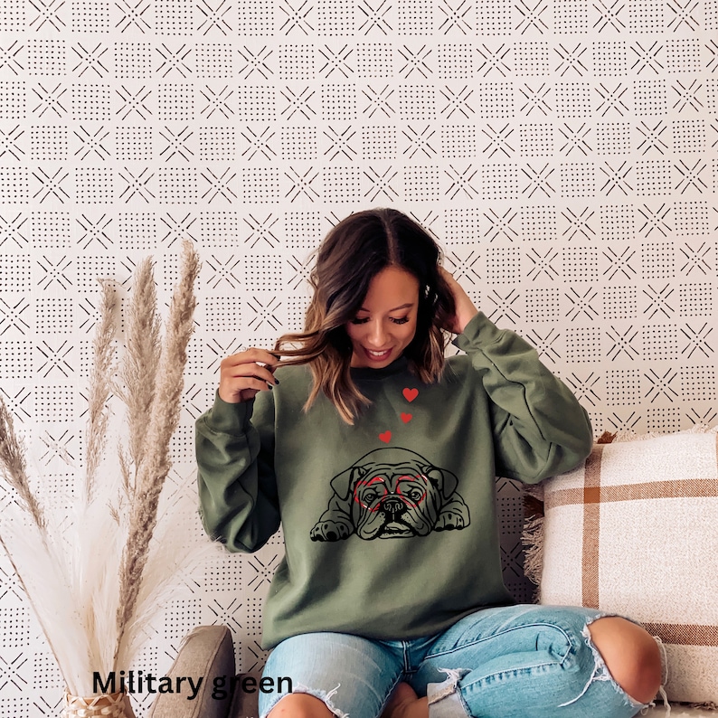 May include: A green sweatshirt with a black bulldog wearing red glasses and surrounded by red hearts. The sweatshirt is on a person wearing blue jeans.