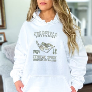 May include: White hooded sweatshirt with a graphic design of a snowboarder in a green and white color scheme. The text on the sweatshirt reads "Freestyle Snowboarding", "Winter Sport", "1995", "Extreme Sport", and "Mountain Are Calling".