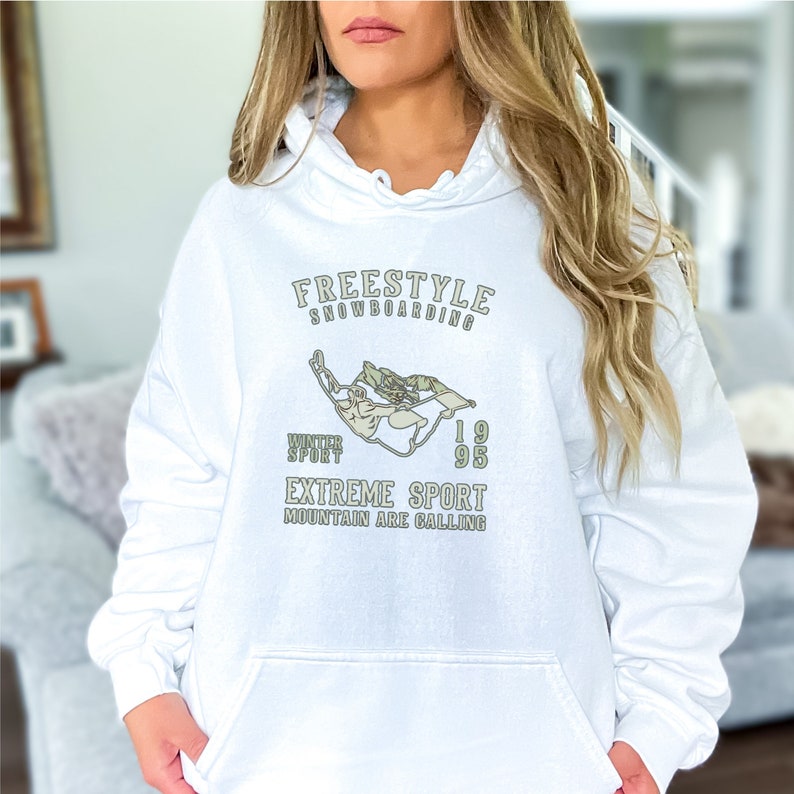 May include: White hooded sweatshirt with a graphic design of a snowboarder in a green and white color scheme. The text on the sweatshirt reads "Freestyle Snowboarding", "Winter Sport", "1995", "Extreme Sport", and "Mountain Are Calling".