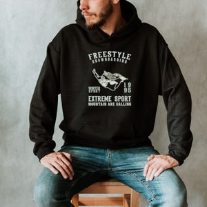 May include: A black hoodie with a white graphic print of a snowboarder. The text on the hoodie reads "Freestyle Snowboarding", "Winter Sport", "1995", "Extreme Sport", and "Mountain Are Calling".