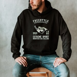 May include: A black hoodie with a white graphic print of a snowboarder. The text on the hoodie reads "Freestyle Snowboarding", "Winter Sport", "1995", "Extreme Sport", and "Mountain Are Calling".