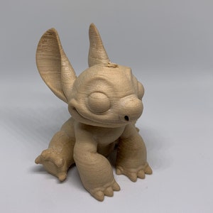 May include: A light brown, 3D-printed figurine of the character Stitch from the animated film Lilo & Stitch. The figurine is in a seated position with large ears, a wide smile, and detailed paws. The figure is made of a light brown material.