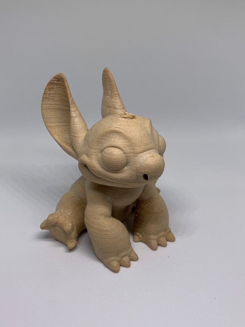 May include: A light brown, 3D-printed figurine of the character Stitch from the animated film Lilo & Stitch. The figurine is in a seated position with large ears, a wide smile, and detailed paws. The figure is made of a light brown material.