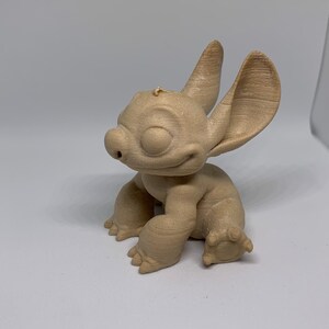 May include: A tan-colored figurine of a cartoon character with large ears, a small nose, and a smiling expression. The figurine is in a seated position with its arms and legs visible. The character's features are detailed, and the overall design is smooth.