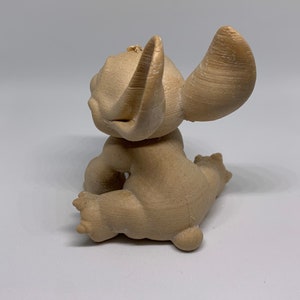 May include: A tan-colored figurine of a cartoon character, seen from the back. The figure has large ears, a small tail, and is in a seated position. The figurine is made of a textured material.