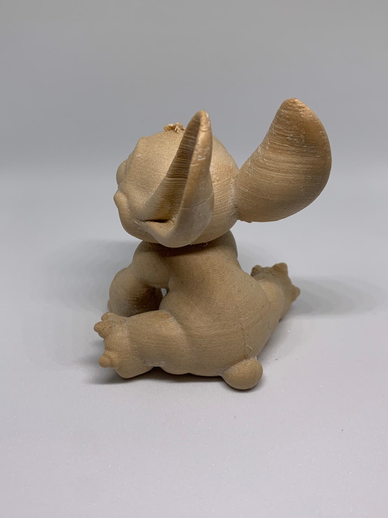 May include: A tan-colored figurine of a cartoon character, seen from the back. The figure has large ears, a small tail, and is in a seated position. The figurine is made of a textured material.