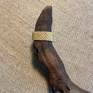 May include: A gold-colored ring with a textured, woven pattern. The ring is displayed on a piece of dark brown wood against a neutral background. The ring's design features a series of small, rectangular blocks, creating a unique and eye-catching texture.