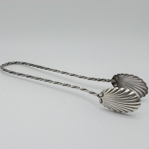 May include: Silver-toned serving tongs with a twisted handle and shell-shaped ends. The tongs are designed for serving and have a decorative, elegant appearance. The shell-shaped ends add a unique touch to the serving utensil.