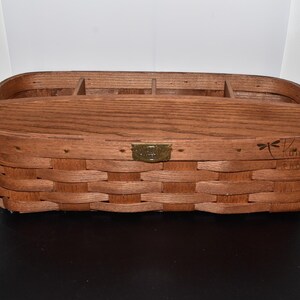 May include: A rectangular, woven wooden basket with a divided interior. The basket is a warm brown color and features a brass-colored metal plate on the front. A dragonfly and text are visible on the side.