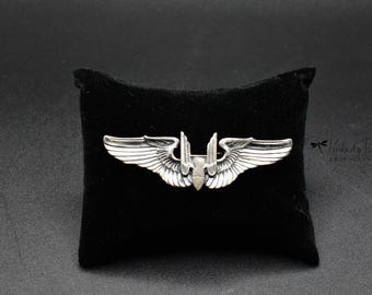 May include: A silver-colored wing pin with detailed feathering, displayed on a black velvet cushion. The pin features a central design with two wings extending outwards. The background is a solid black.