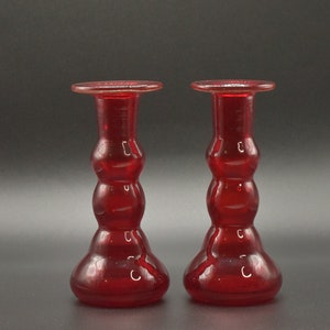 May include: Two red glass candle holders with a bulbous design. The holders have a wide base and a narrow neck.