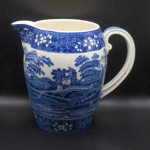 May include: A blue and white ceramic pitcher with a landscape design. The pitcher features a detailed scene of a bridge, trees, and buildings. The handle and rim are white, and the top border has a floral pattern. The pitcher is a vintage piece.