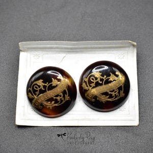 May include: Two round, dark brown buttons with a gold floral design and a gold banner with text. The buttons are on a white paper card with a decorative edge. The buttons have a tortoiseshell pattern.
