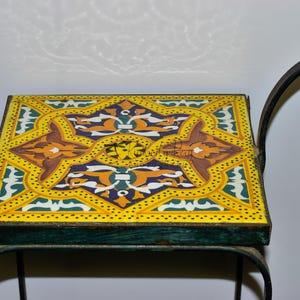 May include: A square, decorative side table with a colorful, patterned tile top. The tile features a yellow background with intricate designs in brown, orange, blue, and green. The table has a dark green metal frame and a curved, wrought iron side.