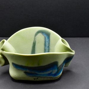 May include: A light green ceramic bowl with a semi-circular back panel and two small tubular side attachments. The bowl features abstract blue and dark grey brushstroke patterns and a small black dragonfly icon printed near the base.