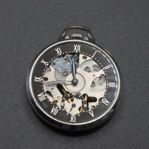 May include: A black and silver pocket watch with a skeleton dial, showcasing the intricate inner workings. The watch features Roman numeral hour markers and delicate hands. The watch is approximately 2 inches in diameter.