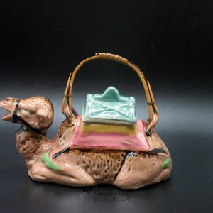 May include: A ceramic camel teapot with a brown, pink, green, and blue glaze. The teapot has a bamboo handle and a lid that looks like a saddle.