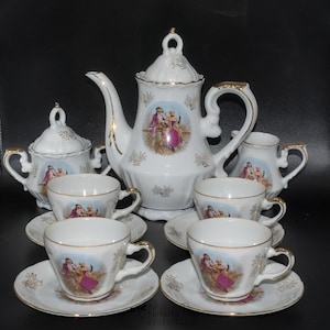 May include: A white porcelain tea set with gold accents and floral designs. The set includes a teapot, creamer, sugar bowl, and four teacups and saucers. Each piece features a romantic couple in a pastoral setting.