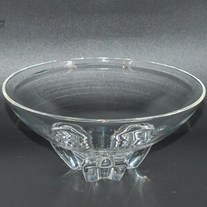 May include: A clear glass bowl with a wide, shallow base and a flared rim. The bowl sits on three small, clear glass feet.