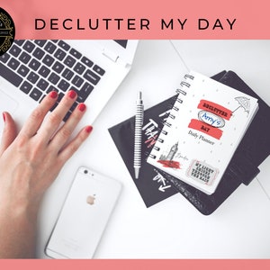 Könnte beinhalten: Ein weißer Tagesplaner mit dem Text "Declutter Amy's Day" und einer Schwarzweißillustration eines Regenschirms und Regentropfen. Der Planer liegt auf einem schwarzen Notizbuch mit einem Aufkleber, auf dem "My light shines through the rain" steht.