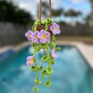 Op de afbeelding: Een gehaakte hangende plantenpot met paarse bloemen en groene bladeren. De pot hangt aan een bruin koord.