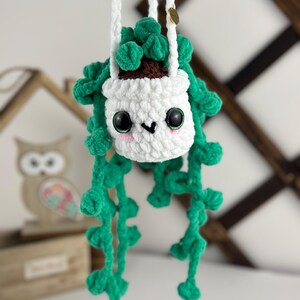 May include: A crocheted hanging planter with a white pot and green leaves. The planter has a cute face with black eyes and a pink nose.