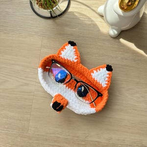 May include: Orange and white crocheted fox-shaped glasses holder with black eyes and a pair of glasses inside.