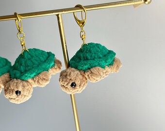May include: Two green and brown crocheted turtle keychains hanging from gold key rings on a gold stand.