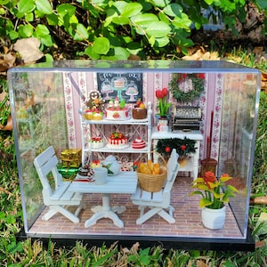 Puede incluir: Una escena de casa de muñecas en miniatura con una mesa y sillas blancas, un estante blanco con pasteles y postres en miniatura, una pequeña corona de Navidad y una planta en maceta. La escena está encerrada en una caja de acrílico transparente.